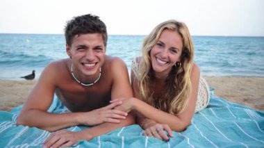 Neşeli bir çift kumsalda güneşli bir günün tadını çıkarırken, renkli bir havluda uzanırken, etrafları sakin deniz ve güneşli mavi gökyüzünün altında yumuşak kumla çevrili kahkahalar ve gülücükler atıyorlar. Yüksek