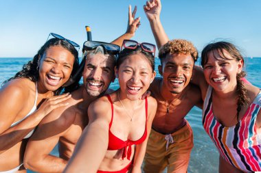 Bir grup neşeli arkadaş sahilde harika vakit geçiriyorlar, gülümsüyorlar ve neşeli bir hatırayı yakalıyorlar parlak ve güneşli gökyüzünün altında ılık deniz suyunda su sıçratıyorlar..