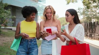 Keyifli bir alışveriş deneyimi yaşayan, cep telefonlarıyla mutlu bir şekilde etkileşime geçen ve alışveriş torbalarıyla birlikte yürürken neşeli anları paylaşan üç kadın..