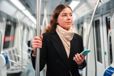 Genç bir kadın metroda seyahat ederken akıllı telefonunu kullanmakla meşgul, bir eliyle direği tutuyor ve diğer eliyle çeşitli uygulamalara bakıyor, tamamen cep telefonuna dalmış.