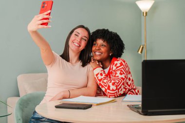 İki neşeli kadın cep telefonuyla eğlenceli bir selfie çekerken masada gülücüklerini ve kahkahalarını paylaşıyorlar, arkadaşlıklarını ve neşeli ilişkilerini kapalı bir ortamda sergiliyorlar.