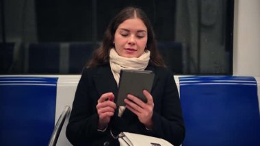 Neşeli genç bir kadın metroda otururken e-okuyucusuna dalmış, seyahati sırasında dijital kitabından keyif alıyor. Yüzündeki ifade, okumayla ilgili bir zevk ve bağlılık olduğunu gösteriyor.