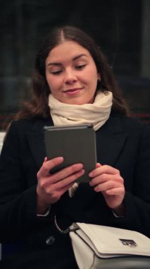 Neşeli genç bir kadın metroda otururken e-okuyucusuna dalmış, seyahati sırasında dijital kitabından keyif alıyor. Yüzündeki ifade, okumayla ilgili bir zevk ve bağlılık olduğunu gösteriyor.