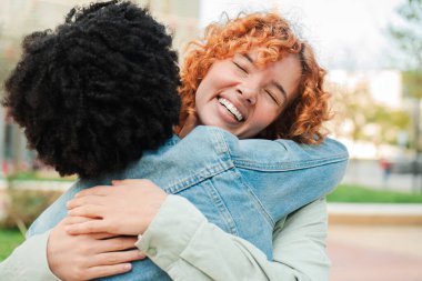 İki kadın birbirlerine sevinçle sarılıyor, şefkatli kucaklaşmalarında mutluluk ve sıcaklığı ifade ediyorlar. Gülüşleri sevgi ve dostluk saçıyor, güzel bir bağın özünü yakalıyor.