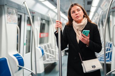 İnterneti taramak için akıllı telefonunu kullanırken direğe tutunan ve günlük rutin işi sırasında metroda mobil bir uygulamayla uğraşan kadın yolcu.