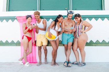 Bir grup arkadaş, güneşli bir sahil gününde akıllı telefonlarıyla keyifli anlar ve kahkahalar atarak neşeli plaj aksesuarları ve yaz havalarıyla çevrelenmiş..