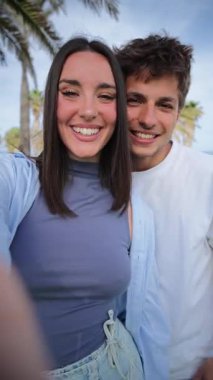 Dikey. Neşeli bir çift gülümsüyor ve eğlenceli bir selfie için poz veriyor, çevrelerinde palmiye ağaçları ile birlikte güzel bir açık havada mutlu anlarını yakalıyorlar..