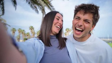 Neşeli bir çift gülümsüyor ve eğlenceli bir selfie için poz veriyor, çevrelerinde palmiye ağaçları ile birlikte güzel bir açık havada mutlu anlarını yakalıyorlar..