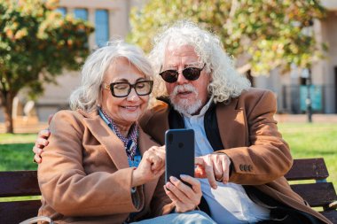 Keyifli, yaşlı bir çift, mutlu bir şekilde akıllı telefon deneyimini paylaşıyor. Uygulamaları incelerken kahkahalara ve sohbetlere dalıp, keyifli bağlarını ve teknolojinin tadını çıkarıyorlar.