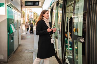 Metro istasyonunun girişinde bekleyen bir bayan yolcu, telefonuna bakarken, aracının gelişini sabırsızlıkla bekliyor, yoğun şehir yaşamındaki bir sonraki macerasına atılmaya hazır..