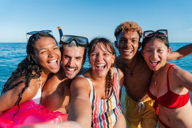 Neşeli bir grup arkadaş, deniz kenarında güneşli bir günde sahilde keyifli bir selfie çeker. Mutluluklarını ve eğlencelerini sergilerken ferahlatıcı su ve güneşin tadını çıkarırlar..