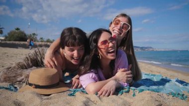 Kumsalda güneşli bir günün tadını çıkaran üç arkadaştan oluşan canlı bir grup. Etrafı kum ve suyla çevrili neşeli bir selfie çekerken gülücükler ve neşe dolu unutulmaz yaz anıları yaratıyorlar.