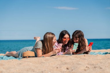 A delightful moment shared by three young women as they explore their smartphones while lying on the warm sand at the beach. Enjoying their time together, they are captivated by their mobile devices.