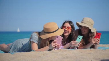 Sahilde sıcak kumda uzanan akıllı telefonlarını keşfederken üç genç kadının paylaştığı keyifli bir an. Birlikte geçirdikleri zamanın tadını çıkarırken, mobil cihazlarıyla büyüleniyorlar..