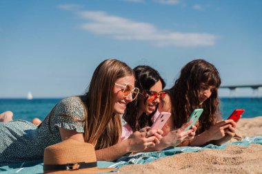Üç mutlu genç kadın kumsalda uzanmış, akıllı telefonlarıyla meşguller, kahkahalarını paylaşıyorlar ve mobil uygulamalar ve mesajlar kullanarak birlikte vakit geçiriyorlar..