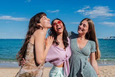 Üç neşeli genç bayan güneşli bir sahilde birlikte harika vakit geçiriyorlar, gülüyorlar ve güzel okyanus manzarasının tadını çıkarıyorlar. Mutlulukları ve dostlukları açık mavi gökyüzünün altında parlıyor..