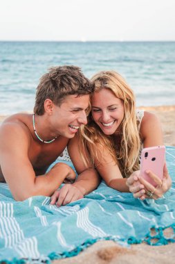 Neşeli bir çift birlikte heyecanlı bir selfie anı yakalıyor, gülüyorlar ve akıllı telefonlarıyla kumsalda eğleniyorlar. Paylaştıkları sevinç bir cep telefonuna bağlandıklarında yayılıyor..