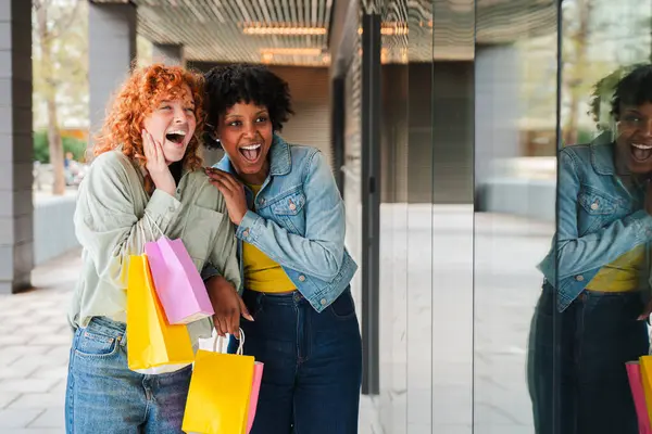 İki neşeli arkadaş başarılı alışveriş gezilerini kutluyor, ellerinde renkli alışveriş torbalarıyla gülüyor ve eğleniyorlar, neşe ve sevinç saçıyorlar.