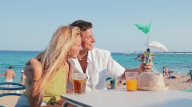 Neşeli çift, güneşli bir sahil gününde, canlı bir barda içkilerini yudumlarken, yaz tatillerinin ve güzel bir sahil manzarasında aşkın özünü sergilerken keyifli bir anı yakalar..