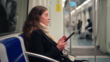 Bir kadın, bir toplu taşıma aracında otururken dijital bir cihaza bağlanıyor, konsantrasyon ve keyif gösteriyor. Sahne, modern banliyö ve kişisel seyahatin özünü yansıtıyor.