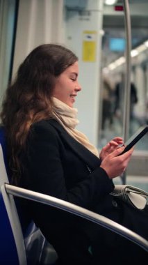 Bir kadın, bir toplu taşıma aracında otururken dijital bir cihaza bağlanıyor, konsantrasyon ve keyif gösteriyor. Sahne, modern banliyö ve kişisel seyahatin özünü yansıtıyor.