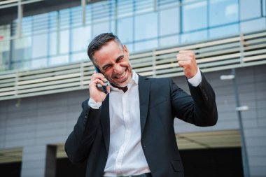 Başarılı bir telefon görüşmesinin ardından heyecanını ifade eden neşeli bir işadamı profesyonel zaferini ve kararlılığını iyi tasarlanmış bir takım elbise içinde sergiliyor. Kendine güveni ve başarısını somutlaştırıyor..