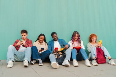 Neşeli bir grup genç arkadaş mobil cihazlarıyla etkileşimde, yerde otururken anları ve kahkahaları paylaşıyor, modern bir bağlantı ve arkadaşlık tarzı sergiliyorlar..