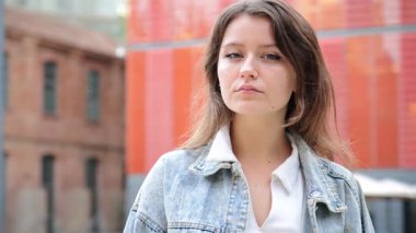 Dışarıda kameraya bakan genç bir kadın, sıradan bir kot ceket giyiyor. Kendine güvenen ve ciddi bir ifade sergileyen genç bir kız doğal ışıkla kentsel bir çevrede kameraya bakıyor. Yüksek