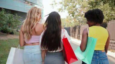 Eğlenceli bir alışveriş deneyimi yaşayan üç neşeli genç bayandan oluşan canlı bir grup ellerinde renkli alışveriş torbalarıyla birlikte yürüyorlar ve gülümsemelerini paylaşıyorlar. Dostluk ışıl ışıl parlar.