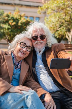 Neşeli yaşlı bir çift, akıllı telefonlarıyla hoş bir selfie çeker ve anı kucaklarlar. Bu iç açıcı sahne, kalıcı anılar oluştururken sevgi ve bağları gözler önüne seriyor.