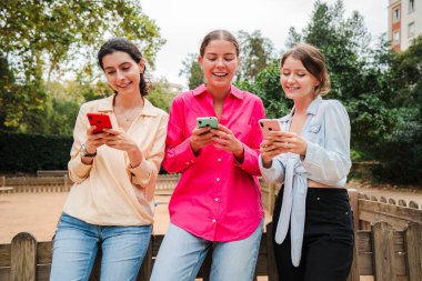 Üç neşeli kadın, etraflarındaki arkadaş ve ailelerle iletişim kurmak ve iletişim kurmak için mobil uygulamalar kullanırken akıllı telefonlarını taramakla, kahkaha ve keyif paylaşmakla meşguller..