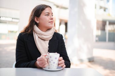 Çekici genç bayan, dışarıda bir teras kafede nefis bir fincan kahve yudumluyor, şık bir ceket ve sıcak bir eşarp giyiyor, yumuşak güneş ışığı altında sakin düşüncelerde kayboluyor.