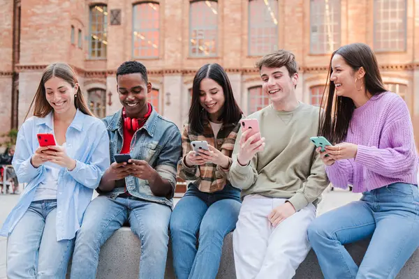 Neşeli bir arkadaş grubu akıllı telefonlarıyla neşe içinde eğleniyor, kahkahalar atıyor ve iletişim anlarını paylaşıyorlar. İfadeleri, gençliğin bu sosyal ortamında mutluluk ve neşeyi yansıtıyor..