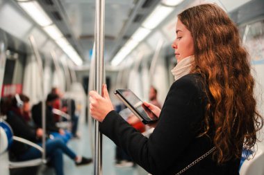 Meşgul kadın, metroda dikilirken e-okuyucusuna odaklandı. Dijital içeriğe tamamen dalmıştı. Şehirdeki günlük ulaşım yolculuğu sırasında destek için bir direğe tutunmuştu.