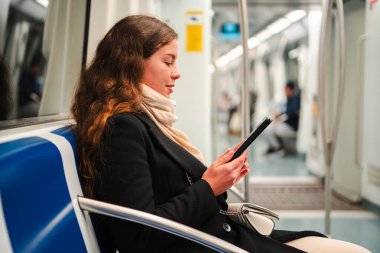 Bir kadın, bir toplu taşıma aracında otururken dijital bir cihaza bağlanıyor, konsantrasyon ve keyif gösteriyor. Sahne, modern banliyö ve kişisel seyahatin özünü yansıtıyor.