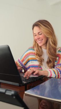 Dizüstü bilgisayarında yazarken renkli bir kazak giyen neşeli bir kadın, verimli ve çekici bir çalışma ortamını iyi ışıklandırılmış bir ofis alanında gösteriyor, kararlılık ve odaklanma gösteriyor..