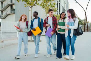 Birlikte eğlenen, gülümseyen ve kahkahalarını paylaşan bir grup genç öğrenci kampüste yürürken canlı dosyalar ve defterler taşıyorlar, dostluk ve dostluk sergiliyorlar..