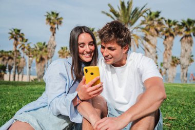 Akıllı telefonlarıyla etkileşime giren, neşeli anları paylaşan ve açık havada güneşli bir ortamda birlikte otururken kahkahalar atan mutlu çiftler. Bağlantı ve sevinç göstermek için mükemmel.