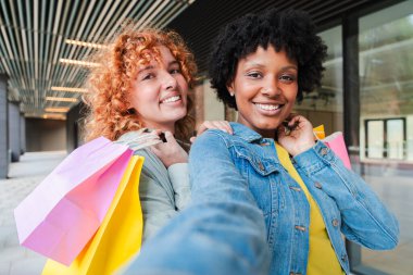 İki neşeli kız heyecanlı bir şekilde alışveriş maceralarını paylaşırken renkli çantalar ve ışıl ışıl gülücüklerle eğlenceli bir selfie çekiyorlar, birlikte keyifli bir anda dostluk ve keyif gösteriyorlar..