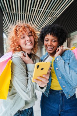 İki neşeli genç kadın, yanlarında renkli çantalarla alışveriş deneyimlerinin tadını çıkarırken mutlu bir şekilde akıllı telefonlarına bakıyorlar ve keyifli ve çekici bir an yaratıyorlar..