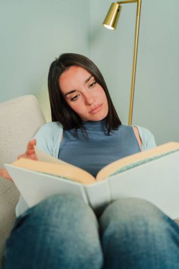 Büyüleyici bir kitaptan zevk alan odaklanmış bir dişi, rahat bir kanepede dinlenirken, edebi keşiflerin heyecanı ve okumanın huzur veren zevkini somutlaştırıyor.