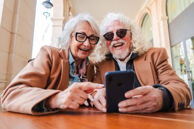 Uygulamaları keşfetmek için akıllı telefonu kullanırken neşeli bir bağ kuran mutlu yaşlı çift, açık havada keyifli bir anın tadını çıkarırken kahkaha ve gülümsemelerini paylaşıyor.