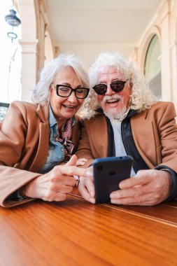 Uygulamaları keşfetmek için akıllı telefonu kullanırken neşeli bir bağ kuran mutlu yaşlı çift, açık havada keyifli bir anın tadını çıkarırken kahkaha ve gülümsemelerini paylaşıyor.