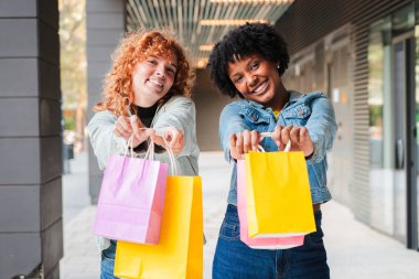 İki hevesli arkadaş, son zamanlardaki alışverişlerini parlak, renkli çantalarla göstererek arkadaşlığın ve perakende terapinin neşesini ve anı paylaşmanın keyifli deneyimini somutlaştırıyorlar..