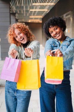 İki neşeli arkadaş keyifli alışveriş deneyimlerini kutlarken ellerinde neşeli mutluluk ve heyecan ifadeleri olan renkli çantalar tutuyorlar.