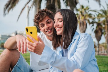 Neşeli bir çift birlikte oturup, eğlenceli bir uygulamayı keşfetmek için akıllı telefonu kullanırken keyifli bir anın tadını çıkarıyor, onların bağlantılarını gösteriyor ve teknolojiyle deneyimlerini paylaşıyor..