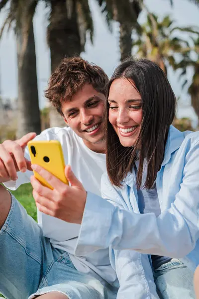 Birbirlerine yakın oturan neşeli çift, akıllı telefonlarıyla meşgul, kahkahalarını ve mutluluk anlarını paylaşırken birlikte güzel bir açık hava ortamında eğleniyorlar..