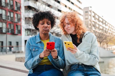 İki kadın birlikte oturuyor, akıllı telefonlarına derinden odaklanmış, cep telefonlarıyla iletişim kurarken endişe ve hayal kırıklığı ifadeleri sergiliyorlar..
