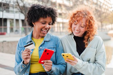 İki neşeli kız cep telefonlarıyla meşgul, kredi kartıyla alışveriş yapıyorlar, ve birlikte hoş bir alışveriş deneyimi yaşarken aynı zamanda gülümseme ve heyecan paylaşıyorlar..
