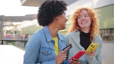 İki neşeli kız cep telefonlarıyla meşgul, kredi kartıyla alışveriş yapıyorlar, ve birlikte hoş bir alışveriş deneyimi yaşarken aynı zamanda gülümseme ve heyecan paylaşıyorlar..